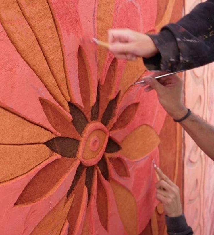 Manos tallan con herramientas una flor de esgrafiado sobre un muro rosa